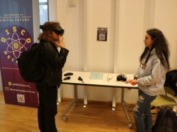 Leila Idrissi stands and watches someone viewing her project through a Virtual Reality headset.